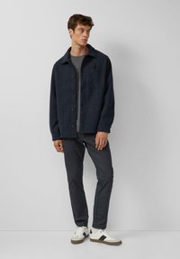 Navy wool-blend button-up jacket with a spread collar, single chest pocket, paired with grey-striped pants and white sneakers with black accents.