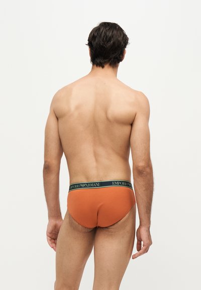 Homme debout de dos portant un slip orange Emporio Armani avec une ceinture noire sur un fond clair uni.