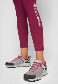 Zapatillas deportivas grises y rosas con superiores texturizados y una suela robusta, combinadas con leggings ajustados burdeos que muestran un logo blanco.