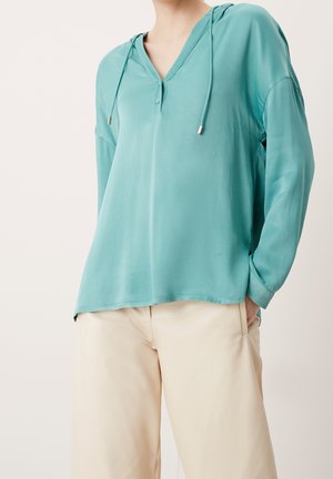 Person wearing a loose teal long-sleeve blouse with a V-neck and drawstrings, paired with beige trousers, standing with one hand in pocket.