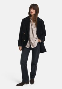 Mulher a usar um casaco preto de dupla-botão, uma camisa de padrão xadrez bege, jeans escuros e botas de cano curto de camurça escuras, de pé com uma mão no bolso.
