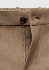 Brown fabric pants close-up, featuring a button closure, zipper, and textured waistband. The material appears smooth with subtle stitching details.