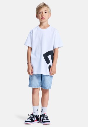 Witte katoenen t-shirt met een grote zwarte afbeelding aan de zijkant, gecombineerd met lichtblauwe denim shorts en zwarte sneakers met rode accenten.