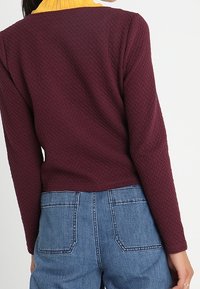 Bordåtsfärgad, texturerad tröja med en ribbad gul krage, med långa ärmar och kort längd, i kombination med högmidjade blå jeans.