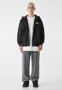 Sort hættejakke med lynlås over en hvid T-shirt, kombineret med grå baggy jeans. Inkluderer sidelommer og en afslappet pasform.