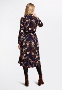 Marinblommig klänning med bälte i midjan, med långa ärmar, rund krage och en svävande, lager på lager kjol. Mörkbruna och gröna toner i mönstret.