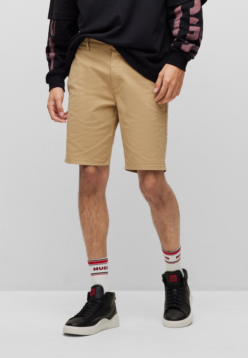 Homme portant un short beige jusqu'aux genoux, des baskets montantes noires, des chaussettes blanches à rayures rouges et noires, et un sweat-shirt noir avec des rayures roses.