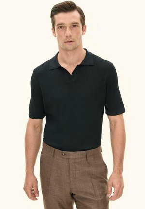 Man wearing short-sleeve black textured polo shirt tucked into brown high-waist trousers, standing against a plain light background.