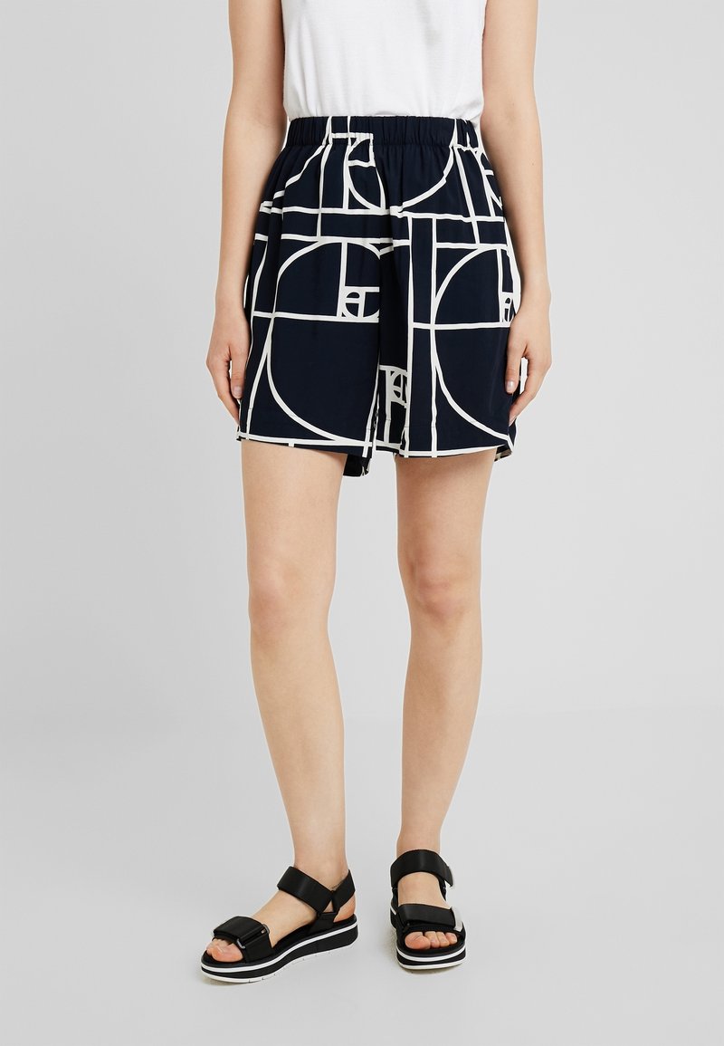 Femme portant un short noir avec des motifs géométriques blancs, un haut blanc sans manches et des sandales à plateforme noires avec des lanières épaisses.