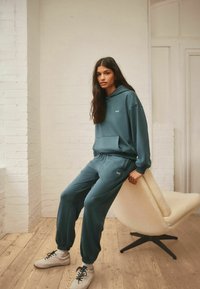 Teal Kapuzenpullover- und Hosen-Set aus einem weichen Material, mit Kängurutasche und elastischen Bündchen. Weißes Logo auf der Brust, kombiniert mit weißen Sneakers.