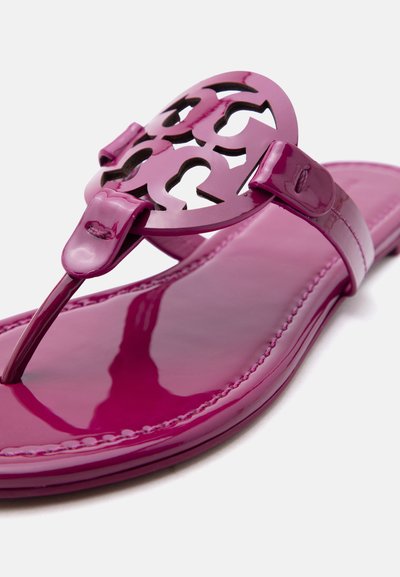 Γυαλιστερό φλατ σανδάλι σε ματζέντα με κομμένο κυκλικό διακοσμητικό έμβλημα και λουράκι ανάμεσα στα δάχτυλα σε απλό φόντο.