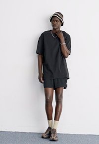 Chemise noire oversize à manches courtes, shorts noirs, chaussettes beiges et baskets brown à motifs. Porté avec un chapeau seau rayé.