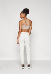 Kvinne som står med ryggen til iført lys beige crisscross sportstopp, hvite joggebukser og svart-hvite joggesko mot en ensfarget bakgrunn.