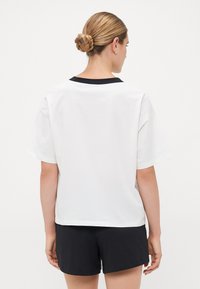 Frau mit blonden Haaren in einem geflochtenen Dutt, die ein weißes Kurzarmshirt mit schwarzem Kragen und schwarze Shorts trägt, steht vor einem weißen Hintergrund.