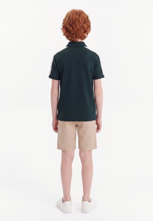 Enfant aux cheveux roux, debout de dos, portant un polo sombre à rayures blanches, un short beige et des baskets blanches sur fond blanc.