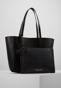 Černá kožená taška přes rameno s dvěma dlouhými uchy, která obsahuje odnímatelnou zipovou kapsu s tmavým vzorem a logem "Calvin Klein" na přední straně.