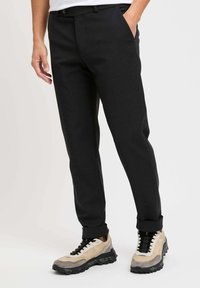 Pantaloni neri su misura a gamba dritta, con tasche laterali e una texture liscia. Abbinati a sneaker beige e nere.