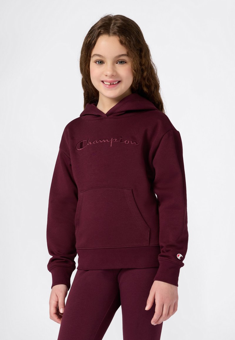 Fille avec de longs cheveux bouclés portant un hoodie maroon Champion et des leggings assortis, souriant et se tenant contre un fond blanc.