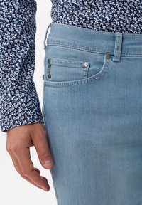 Persona vestida con una camisa de flores azul y jeans azul claro, con una mano relajada al lado de la pierna, mostrando el bolsillo de los jeans y los detalles de la costura.