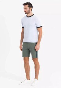 T-shirt bleu clair pour homme avec des bordures noires, associé à un short vert avec des rayures sur les côtés et des baskets blanches. Design simple, coupe décontractée.