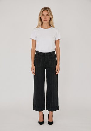 Donna in piedi con il volto rivolto in avanti che indossa una semplice maglietta bianca, pantaloni neri a gamba larga a righe sottili con orli risvoltati e tacchi alti neri.