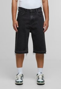 Svarte denimshorts med frynsete kanter, fem-lommers design og en avslappet passform. Kombinert med grønne og hvite joggesko og hvite ankelsokker.