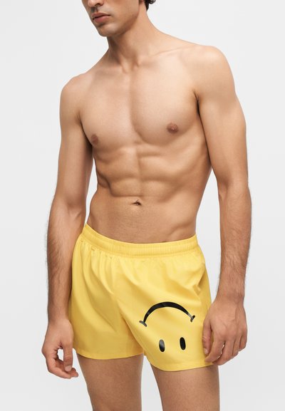 Homme torse nu portant un short jaune avec un grand visage triste noir imprimé sur la cuisse gauche sur un fond uni.