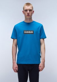 Blå bomulds t-shirt med korte ærmer, rund halsudskæring og et rektangulært logo-patch på brystet, der har sort tekst på hvid baggrund.