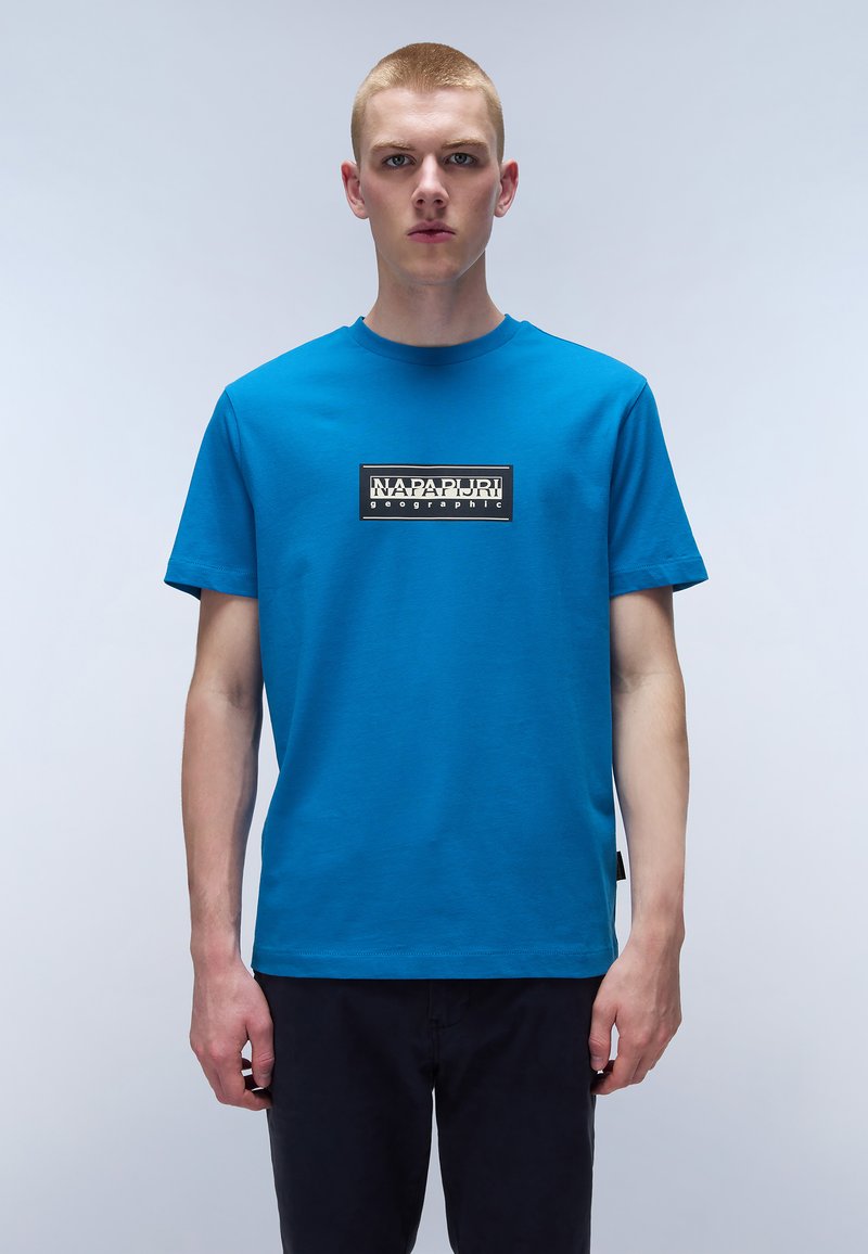 Blå bomulds t-shirt med korte ærmer, rund halsudskæring og et rektangulært logo-patch på brystet, der har sort tekst på hvid baggrund.