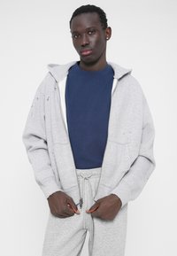 Sweat à capuche gris à fermeture éclair avec intérieur blanc, superposé sur un t-shirt bleu marine. Pantalon de survêtement gris avec cordon de serrage et détails zippés.