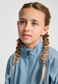 Ljusblå zip-jacka med en texturerad yta, hög krage och två sidoflätar. En liten hoopörhänga bärs i vänster öra.