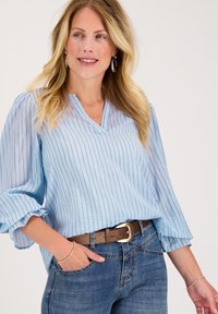 Lichtblauwe blouse met verticale strepen, V-hals en gerimpelde mouwen. Gecombineerd met donkerblauwe jeans en een bruine riem met een gouden gesp.