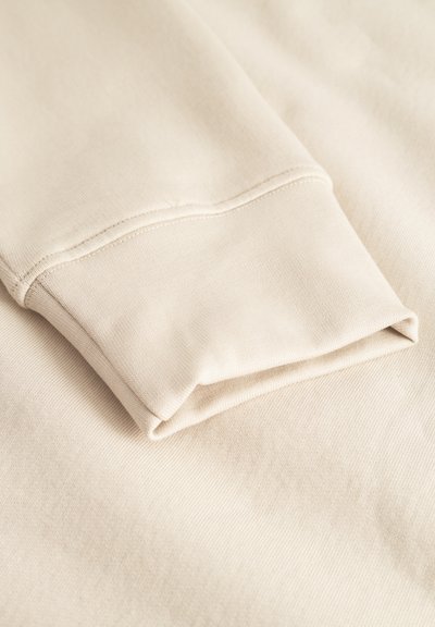 Beiger Sweatshirt-Stoff mit umgeschlagenem Bündchen, der eine glatte Textur und subtile Nähdetails aufweist. Einfaches, funktionales Design.