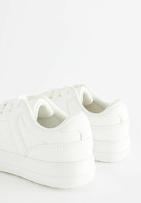Baskets sportives blanches avec une tige synthétique lisse, bout rond, col rembourré et semelle texturée. Présente un design à lacets et des détails de couture.