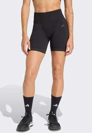 Femeie purtând pantaloni scurți atletici negri cu talie înaltă, șosete negre până la jumătatea gambelor și adidași negri, stând în picioare pe un fundal deschis.