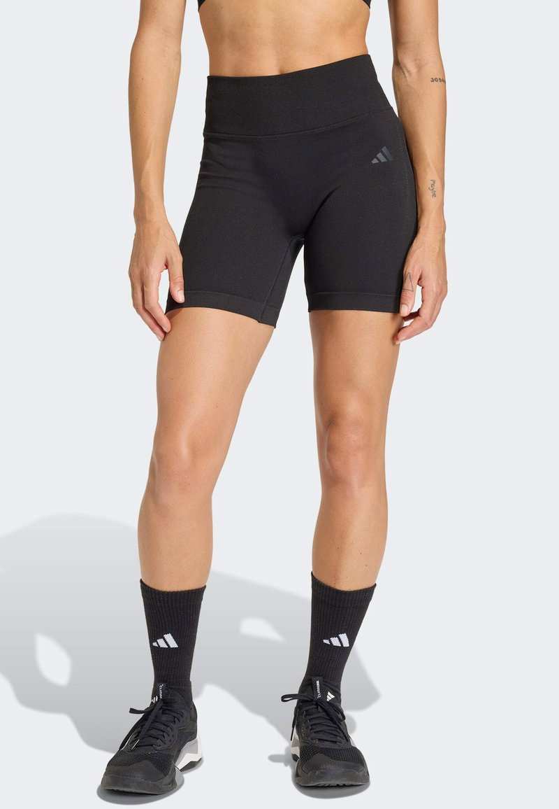 Γυναίκα που φοράει μαύρο αθλητικό σορτς με ψηλή μέση, μαύρες κάλτσες μέχρι τη μέση της γάμπας και μαύρα αθλητικά παπούτσια, στέκεται μπροστά σε ανοιχτό φωτεινό φόντο.