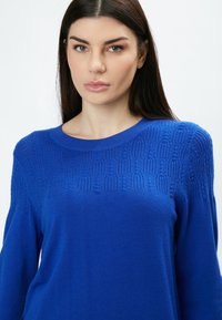 Blauer gestrickter Pullover mit einem strukturierten Design im oberen Bereich, rundem Ausschnitt und langen Ärmeln, aus einem weichen Material gefertigt.