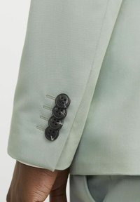 Manga de traje verde claro con una textura suave, que presenta cuatro botones negros y costuras en contraste a lo largo del puño.
