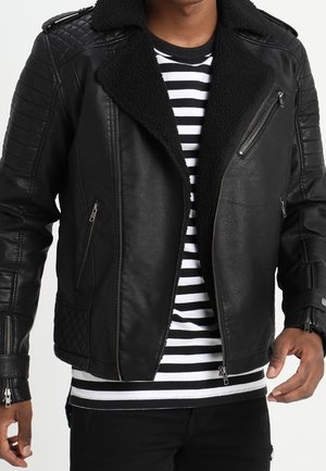 Homme portant une veste de motard en cuir noir avec des panneaux matelassés sur une chemise rayée noir et blanc et un pantalon noir.