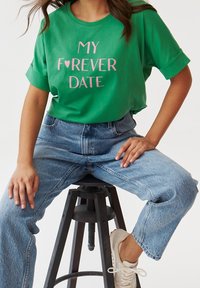 Camiseta de algodón verde con "MI ♥ CITA PARA SIEMPRE" impresa en rosa, con mangas cortas enrolladas y un ajuste holgado, combinada con jeans de tiro alto en azul.