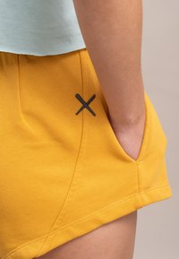 Gele katoenen shorts zijn voorzien van een zijzak en een zwarte geborduurde accent in de vorm van een "X." De stof heeft een gladde textuur.