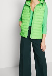 Gilet piumino verde con cappuccio, caratterizzato da cuciture orizzontali e una cerniera frontale. Indossato sopra un top a coste verde abbinato e pantaloni scuri.