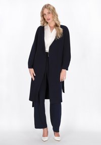 Langer navyblauer Cardigan mit V-Ausschnitt, offener Vorderseite und weit geschnittenen Ärmeln, kombiniert mit einer cremefarbenen Bluse und passenden navyblauen Hosen. Weiße High-Heels.