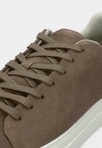 Nærbilde av taupe semsket skinn-sneakers med matchende snøring og hvit såle, som viser detaljert tekstur og søm.