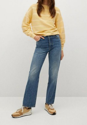 Osoba ubrana w żółty sweter w paski, niebieskie jeansy z wysokim stanem o prostym kroju oraz beżowe trampki, stojąca na tle jednolitego tła.