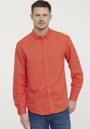 Chemise rouge à manches longues avec boutons, en tissu texturé, dotée d'un col et d'un logo sur la poitrine, associée à un jean en denim bleu.