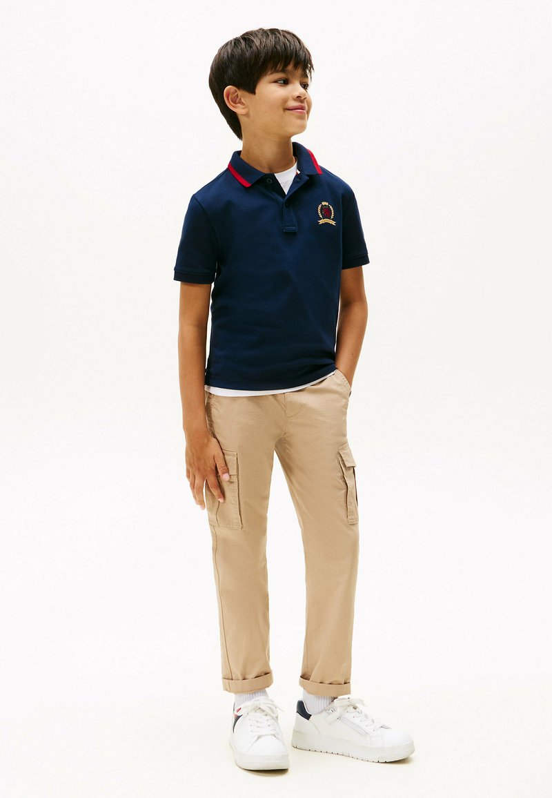 Polo shirt blu navy con colletto rosso e logo ricamato, abbinato a pantaloni cargo beige e sneakers bianche. Tessuto morbido, vestibilità casual.