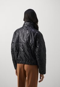 Black puffer jacket featuring a glossy woven texture, elastic hem, and repeating logo pattern. High collar; oversized fit, paired with brown pants.