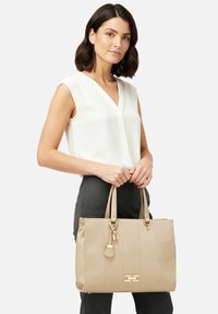 Borsa tote in pelle beige con cuciture verticali, ferramenta dorata e targhetta staccabile. Due manici superiori per il trasporto. Texture liscia.