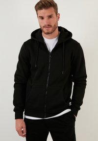 Crna zip-up dukserica od mekanog materijala, s kengur M džepom, kapuljačom s vezicom i rebrastim manžetama. Nosena preko bijele majice.